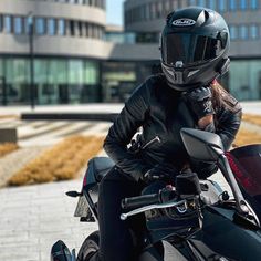 a woman wearing a black leather jacket and helmet sitting on a motorcycle in front of a building