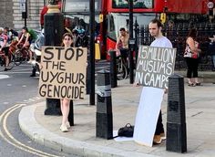 two people holding signs on the sidewalk near a bus stop with other people standing around