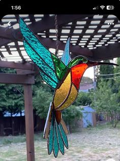 a stained glass hummingbird hanging from a wooden pergolated structure with trees in the background