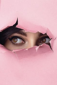 a woman's eye peeks through a hole in the pink paper that has been torn open