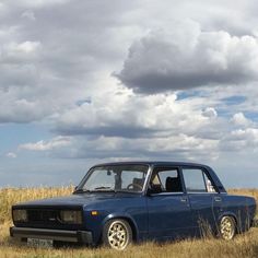 an old car is parked in the middle of a field with tall grass and clouds