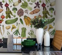 a kitchen counter topped with green plates and vases filled with vegetables next to a wallpaper