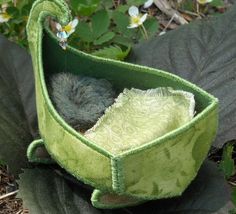 a small stuffed animal in a green fabric bag on the ground next to flowers and leaves