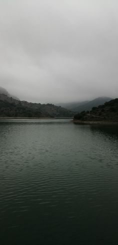 a body of water with hills in the distance on a cloudy day and some clouds