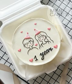 a white cake with pink hearts and two women on it in a plastic container that says 1 year