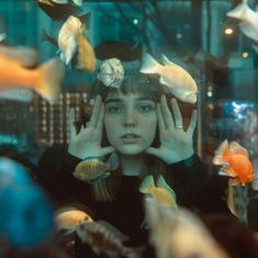 a woman with her hands on her head surrounded by fish in an aquarium looking at the camera