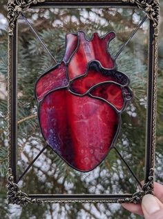 a hand holding up a stained glass heart in front of a pine tree with snow on the branches