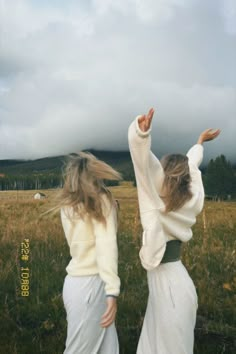 two women in white dresses are walking through a field with their arms spread out to the sky