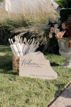 a welcome sign sitting in the grass next to a vase with flowers and candles on it