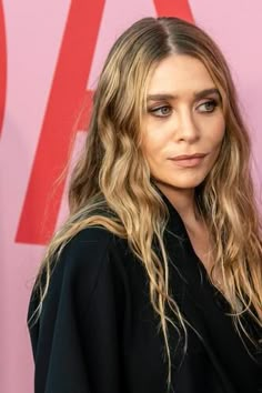 a woman with long blonde hair standing in front of a pink wall and wearing a black top