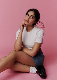 a woman sitting on the ground in front of a pink background wearing white shirt and blue skirt