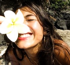 a woman holding a flower up to her face and smiling at the camera while laying on rocks