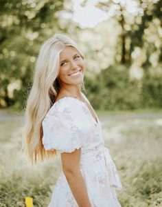 a woman with long blonde hair standing in tall grass smiling at the camera while wearing a white dress