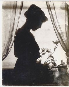 an old black and white photo of a woman sitting in front of a window with flowers