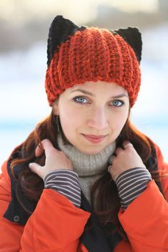 a woman wearing an orange hat with black cat ears on it's head and holding her hands to her ear