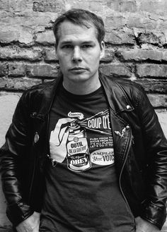 a black and white photo of a man in a leather jacket leaning against a brick wall