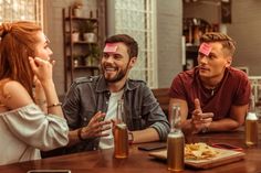 three people sitting at a table having conversation with sticky notes on their foreheads,