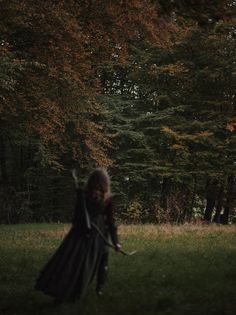 a woman in a long dress holding a bow while standing in the grass with trees behind her