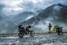 three motorcycles parked on the side of a mountain road with mountains in the background and foggy sky