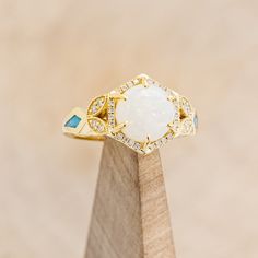 a white opal and diamond ring sitting on top of a wooden stand in front of a beige background