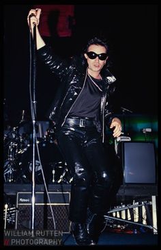 a man in black leather pants and sunglasses on stage holding a microphone with his right hand