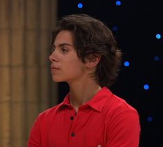 a young man in a red shirt is looking off into the distance with blue lights behind him