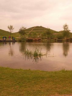 people are standing on the edge of a lake with grass in front of it and hills in the background