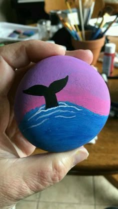 a hand holding a painted rock with a whale tail on it's side in front of a table