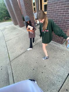 two girls are standing on the sidewalk and one is holding a skateboard in her hand