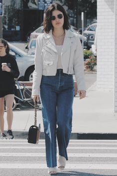 a woman in white jacket and jeans crossing the street with her hand on her hip