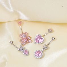 three pairs of pink and white diamond heart shaped belly rings on a satin material background