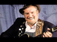 an older man wearing a hat and holding a guitar in his right hand while smiling at the camera