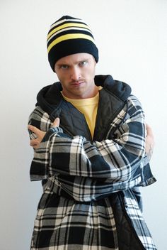 a man wearing a plaid coat and a beanie is standing with his arms crossed