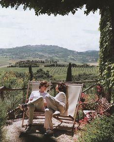 two people sitting on a bench in the middle of an outdoor area with flowers and greenery