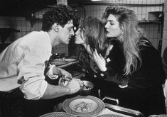 two women and a man standing in a kitchen next to each other with plates on the table