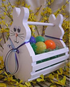 an easter basket with painted eggs in it and a bunny sitting on the top, surrounded by yellow flowers