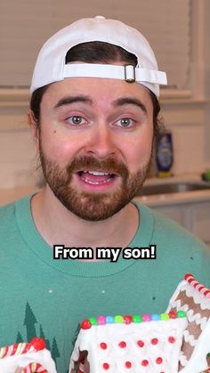 a man wearing a white hat and green shirt holding two gingerbread houses in front of him