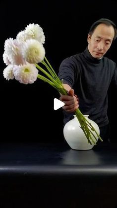 a man arranging flowers in a white vase on a black table with a dark background