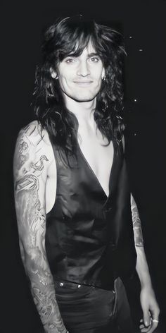 a man with long black hair and tattoos on his arm standing in front of a dark background