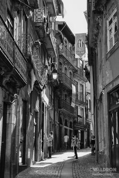 black and white photograph of people walking down an alleyway between two buildings with balconies