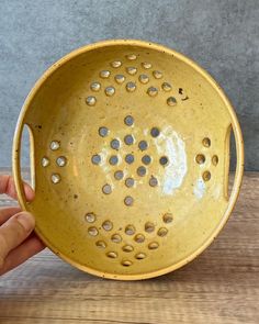 a hand holding a yellow bowl with holes in it on a wooden table next to a gray wall