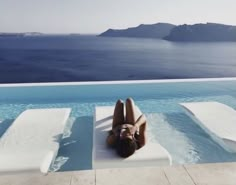 a woman laying on top of a white bed next to a swimming pool with an ocean view