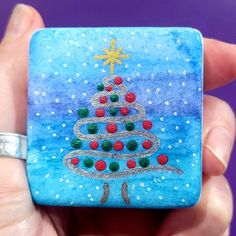 a hand holding a small glass tile with a christmas tree painted on it