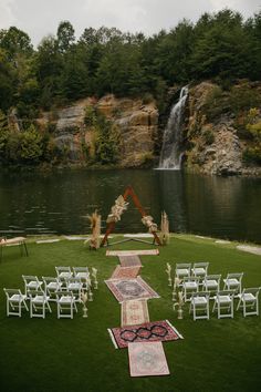 an outdoor ceremony setup with chairs and rugs in front of a waterfall, surrounded by lush green grass