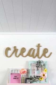 the word create is spelled in gold letters on a white wall above a pink desk