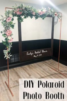 a photo booth with pink flowers and greenery on the wall, in front of a wooden floor