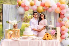 a pregnant couple standing in front of a table with cake and desserts on it