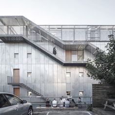 several people are standing in front of a building that has stairs on the side and two cars parked near it