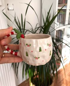 a hand holding a white cup with christmas decorations on it and a green plant in the background