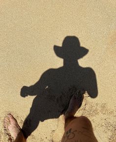 a shadow of a man in a cowboy hat on the beach with his foot up